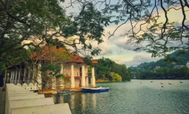 Scenic view of Kandy Lake with traditional pavilion and boats, surrounded by trees and hills in Sri Lanka.