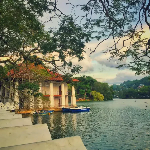 Scenic view of Kandy Lake with traditional pavilion and boats, surrounded by trees and hills in Sri Lanka.