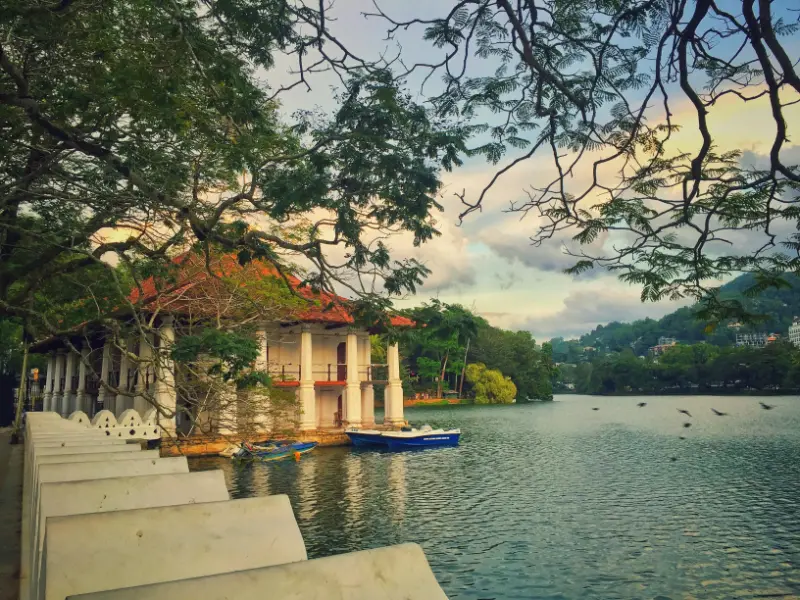 Scenic view of Kandy Lake with traditional pavilion and boats, surrounded by trees and hills in Sri Lanka.